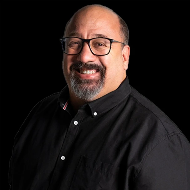 Bald man with glasses and dark shirt smiling against black background, testimonial photo